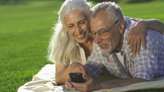 Closeup portrait of happy senior couple lying on green lawn and using smarphone. Elderly man and woman with gray hair using modern device, laughing and enjoying technology while sharing travel photos