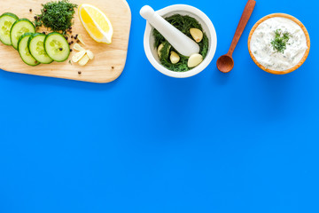 Preparing greek cucumber yogurt sauce. Bowl with yogurt near greenery, cucumber, oranges on cutting board on blue kitchen desk top view copy space