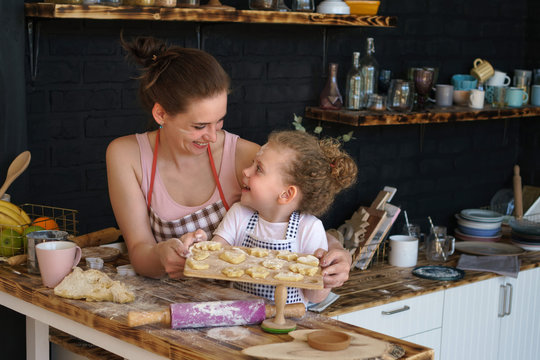 Young mother and daughter prepare cookies in kitchen. They are in aprons. Little girl and woman are holding cutting board with biscuit and looking at each other. Family time.