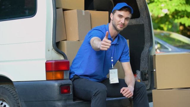 Moving company worker showing thumbs up, sitting in van full cardboard boxes