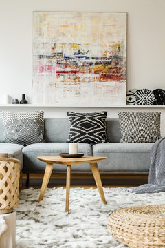Painting above grey couch in boho living room interior with wooden table and pouf. Real photo