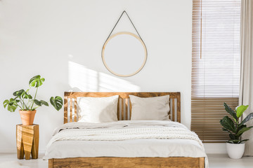 Sun shining on a white wall with a round mirror in a minimalist bedroom interior with natural, wooden furniture and beautiful green plants - obrazy, fototapety, plakaty