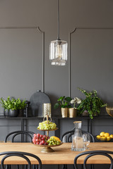 Lamp above wooden table with fruits in grey dining room interior with plants. Real photo