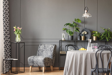 Pink flowers next to grey armchair in flat interior with round dining table under lamp. Real photo
