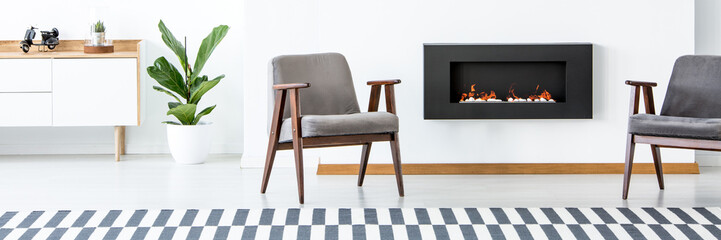 Two grey armchairs standing in white living room interior with fireplace, fresh green plant and cupboard with motorcycle model and cactus