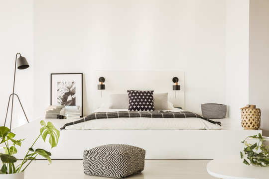 Patterned pouf in front of white bed in bright bedroom interior with plant, lamp and poster. Real photo