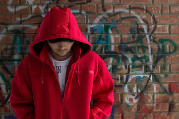 The guy in the red hoodie against the brick wall hides his eyes behind a cap - obrazy, fototapety, plakaty