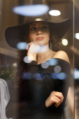 Young beautiful girl with perfect make-up, wearing a black hat and elegant black dress, posing near glass storefront of the shopping center.