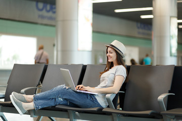 Young calm traveler tourist woman in hat working on laptop while waiting in lobby hall at international airport. Passenger traveling abroad on weekends getaway. Air travel, flight journey concept.