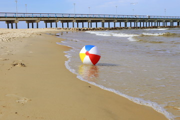 Obraz premium Colorful beach ball on sand and waving sea. Holiday beach ball near Baltic sea and Palanga bridge in horizon.