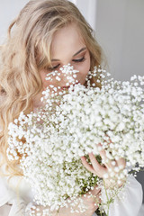 Naklejka premium Curly blonde romantic look, beautiful eyes. White wildflowers in hands. Girl white light dress and curly hair, portrait of woman with flowers at home near the window, purity and innocence