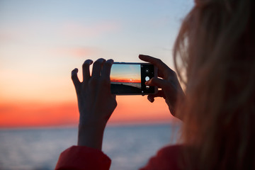 Woman traveler using smartphone and taking photo of colorful sea sunset