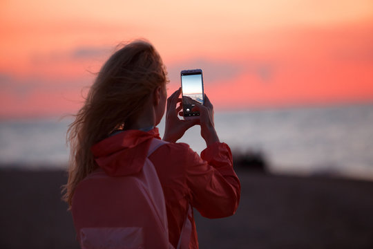 Woman traveler using smartphone and taking photo of colorful sea sunset