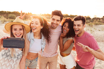 Photo of smiling young multiracial men and women 20s in summer clothes taking selfie on smartphone,...