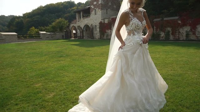 Rear full-length view of the young charming blonde bride in the beautiful long dress running along the blooming garden.