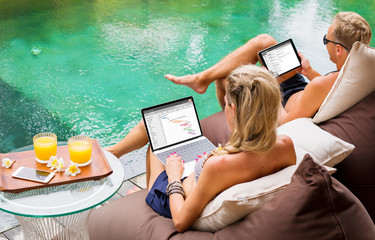 Couple working on computers while relaxing by the pool.