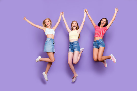 Portrait of crazy funky girls in jeans shorts skirt jumping in air holding raised arms celebrating achievement yelling looking at camera isolated on bright violet background
