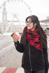 Fototapeta premium Cute young caucasian brunette woman student on a walk in europe city streets. She wearing blac coat and plaid red scarf. Cold winter weather. Woman drinks coffee from a to-go cup