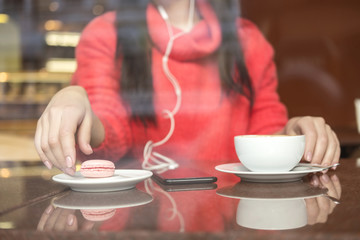 Cute brunette plus size woman working on a distance (freelance), sitting in cafe, drinking coffee and eating macaron. She wearing red sweater, listen music and typing messages on her phone