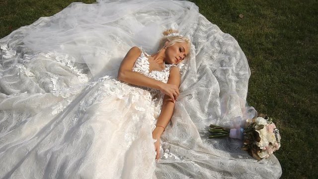 Above view at the young attractive blonde bride lying on the grass and tenderly touching her arm.