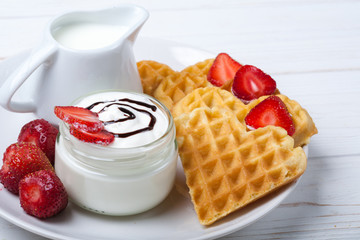 Heart shaped waffles with strawberries, cream sauce, pitcher with milk on white plate and white wooden background. St Valentines day breakfast. Romantic breakfast dessert. Copy space