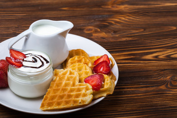 Heart shaped waffles with strawberries, cream sauce, pitcher with milk on white plate and wooden background. St Valentines day breakfast. Romantic breakfast dessert. Copy space