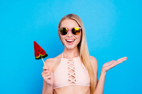 Portrait Of Cheerful Positive Girl In Eyeglasses With Beaming Smile Gesturing With Palm Enjoying Daydream Holding Sweet Tasty Piece Of Watermelon On Stick Isolated On Vivid Blue Background