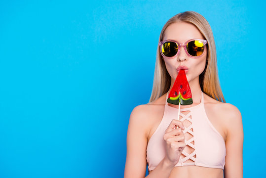 Yummy! Portrait with copy space of attractive charming girl in eyewear eating tasty sweet piece of watermelon on stick wearing pink bra isolated on bright blue background