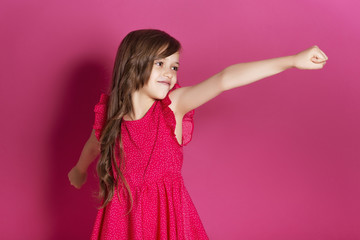 Little 8 years old girl make some emotional gesture with her hands on a pink neutral background. She has long brunette hair and wear red summer dress. Funny expression on her face