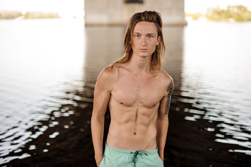 Portrait of young handsome long and red-haired man dressed in shorts standing in water