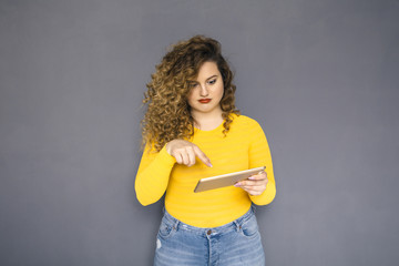 Cute brunette plus size woman with curly hair in yellow sweater and jeans standing on a neutral grey background. She try to use tablet and don't know how to work with it