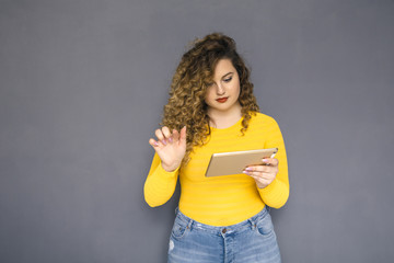 Cute brunette plus size woman with curly hair in yellow sweater and jeans standing on a neutral grey background. She try to use tablet and don't know how to work with it