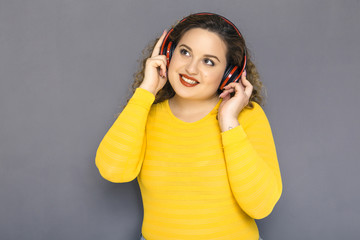 Cute brunette plus size woman with curly hair in yellow sweater and jeans standing on a neutral grey background. She listen music using wireless headphones, and dancing a bit