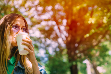 Beautiful young girl drinks take-away cup of coffee in the open air. Fashionable hipster model in heart-shaped sunglasses. Concept of an invigorating drink. Face close-up