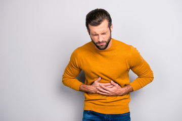 Portrait of sad upset troubled  unhappy ill unhealthy bearded grimacing frustrated guy wearing yellow sweater keeping hands on belly isolated on gray background