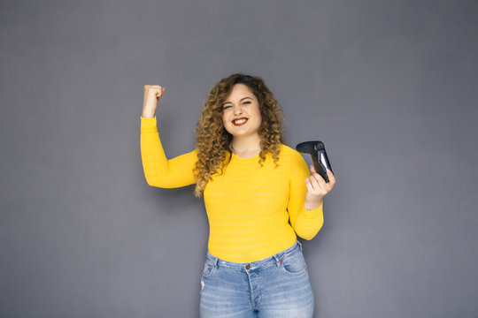 Cute brunette plus size woman with curly hair in yellow sweater and jeans standing on a neutral grey background. She holds a joystick, playing on console games