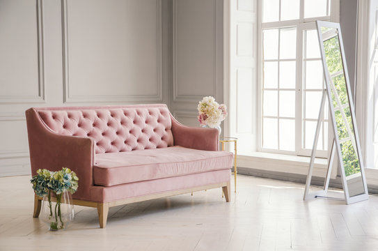 White room with pink sofa-bed, flowers in glass jug and mirror near the window. Classic interior design