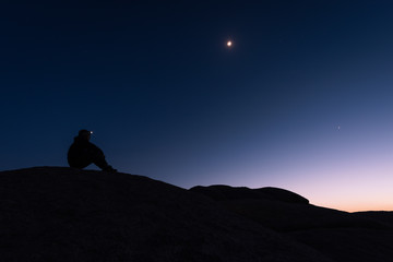 The silhouette of a lone hiker sitting on a rock formation in the desert with a headlamp shining...
