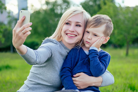 pretty blonde girl (mother) and cute son make selfie on phone in the park on a background of green grass and trees