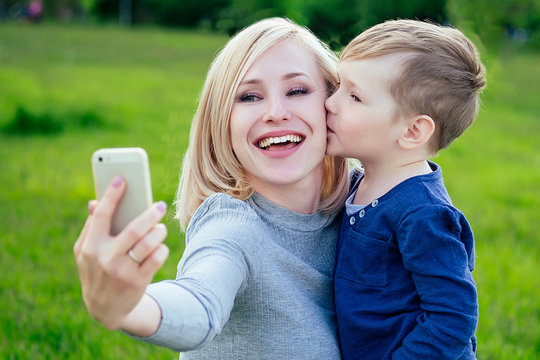 attractive mother (female person) and little boy take pictures selfie on phone in the park on a background of green grass and trees