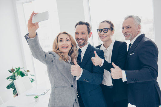 Attractive,  stylish, joyful, confident business persons standing in workplace, workstation, shooting selfie on front camera, using smart phone, having break, fun, showing thumb up with hands, fingers