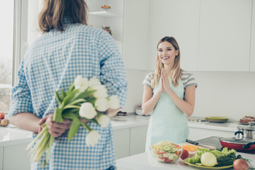Cropped portrait, rear view of handsome man hiding behind back bouquet of white tulips preparing surprise for glad lover in apron standing near table stove making healthy meal