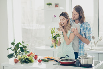 Portrait of lovely romantic couple enjoying valentine day, handsome man with long hair giving white tulips to lover while preparing meal lunch