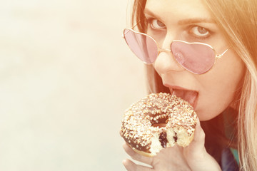 Beautiful young girl stuck out her tongue and tries to lick a donut. Fashionable hipster model in heart-shaped sunglasses. Close up. Mockup