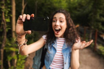 Blurry photo of excited joyful woman 18-20 with backpack, shouting and taking selfie photo on focused black cell phone while walking in green park