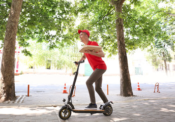 Young man with pizza box riding a kick scooter outdoors. Food delivery service