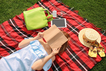 Young woman with book resting on plaid outdoors