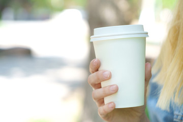 Girl is holding white cup, mug in hands. Mockup for products presentations, logo and text. Hand holding paper cup of coffee in a green park. Branding mockup scene