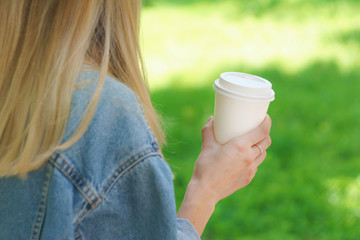 Girl is holding white cup, mug in hands. Mockup for products presentations, logo and text. Hand holding paper cup of coffee in a green park. Branding mockup scene