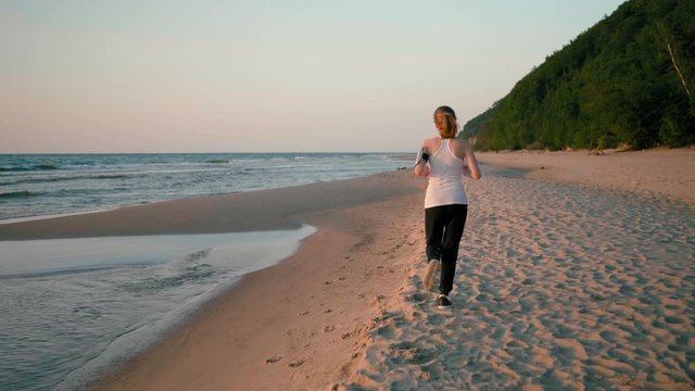 Back View of a Running Sporty Girl Jogging on Beach at the Sea Coast with HeadPhones at Sunset